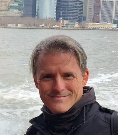 A man taking a selfie on a boat in front of a city skyline with tall buildings and water in the background.