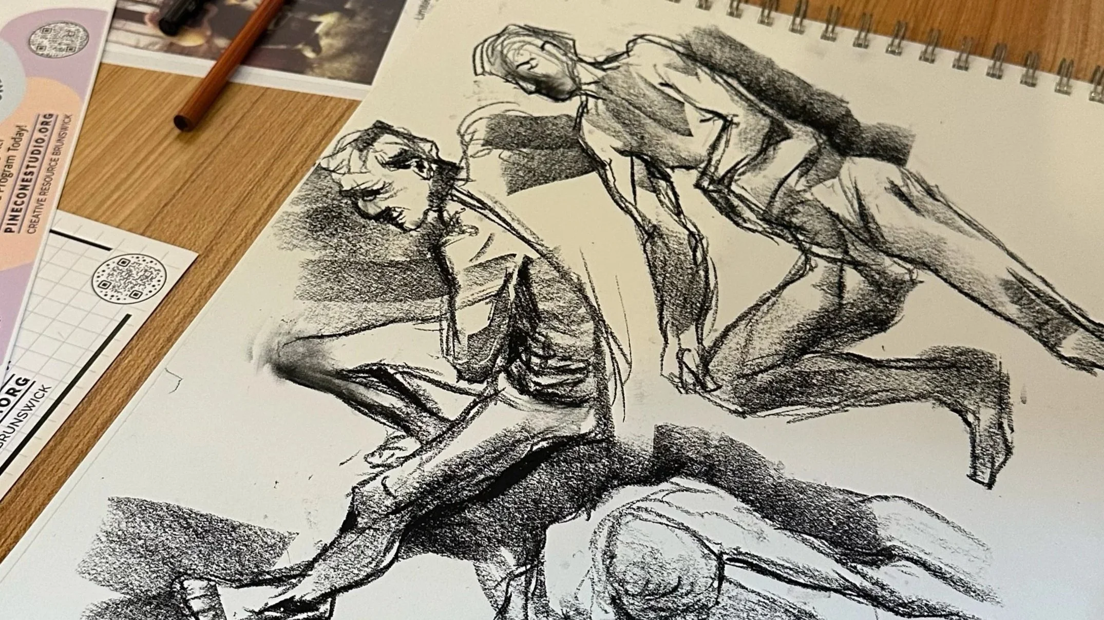 A sketch of three nude human figures on a sheet of paper, appearing to be life drawings or studies, with a wooden table and art supplies around them.