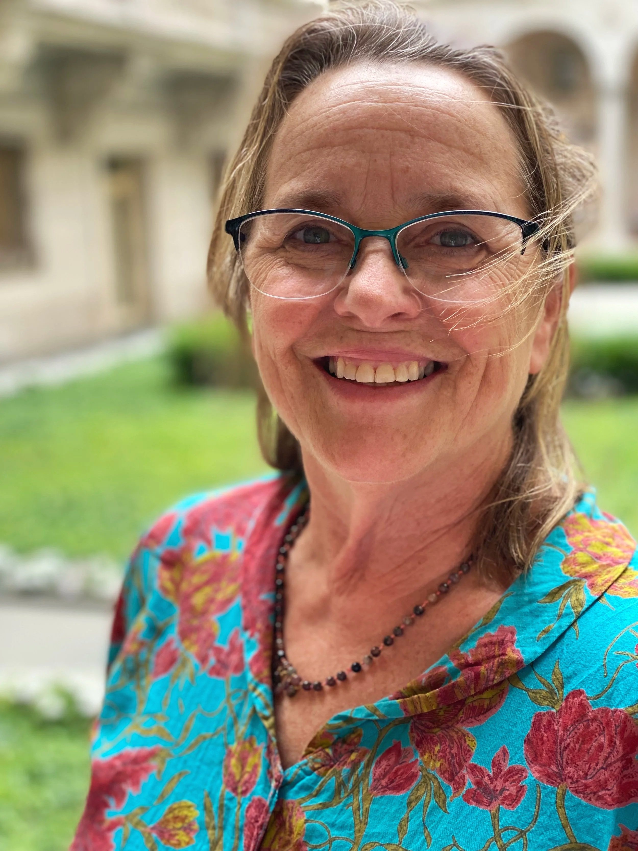 A smiling woman with glasses and shoulder-length hair outdoors in front of a historic building, wearing a colorful floral shirt and a beaded necklace.