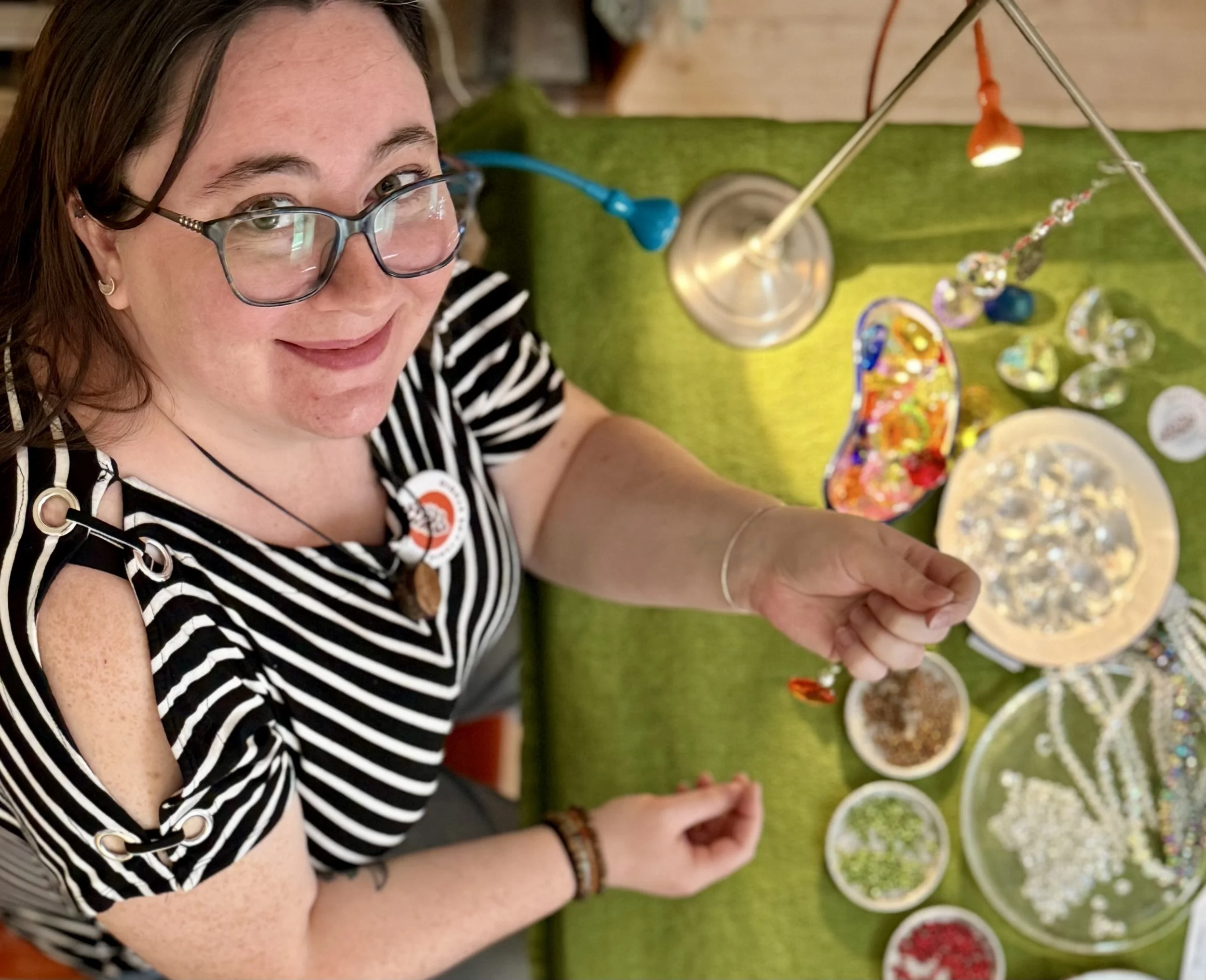 Woman with glasses and a striped shirt smiling while working on a jewelry craft table with beads and jewelry pieces.