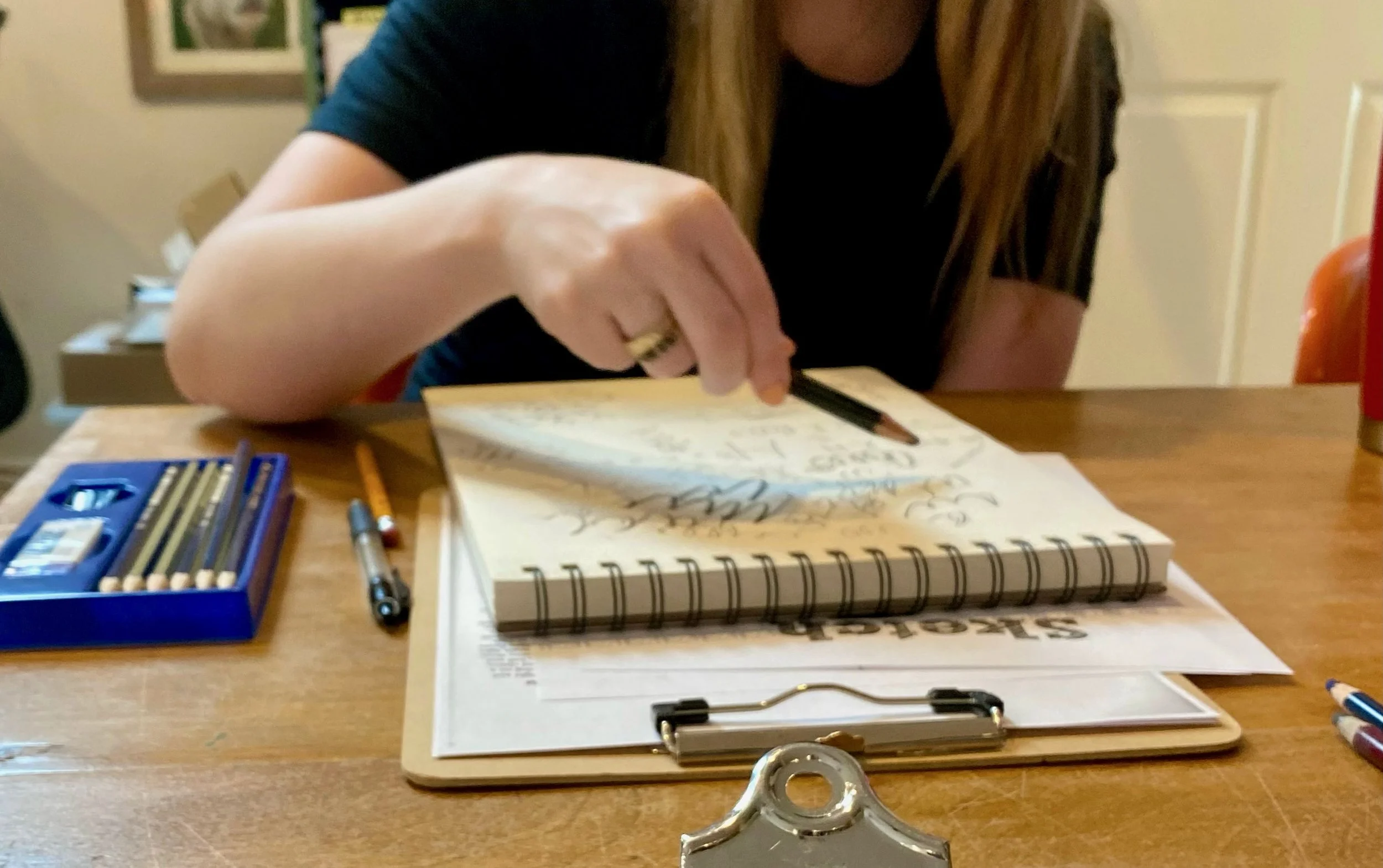 A person with long hair is writing on a spiral notebook with a pen, surrounded by pencils and a paper on a wooden table.