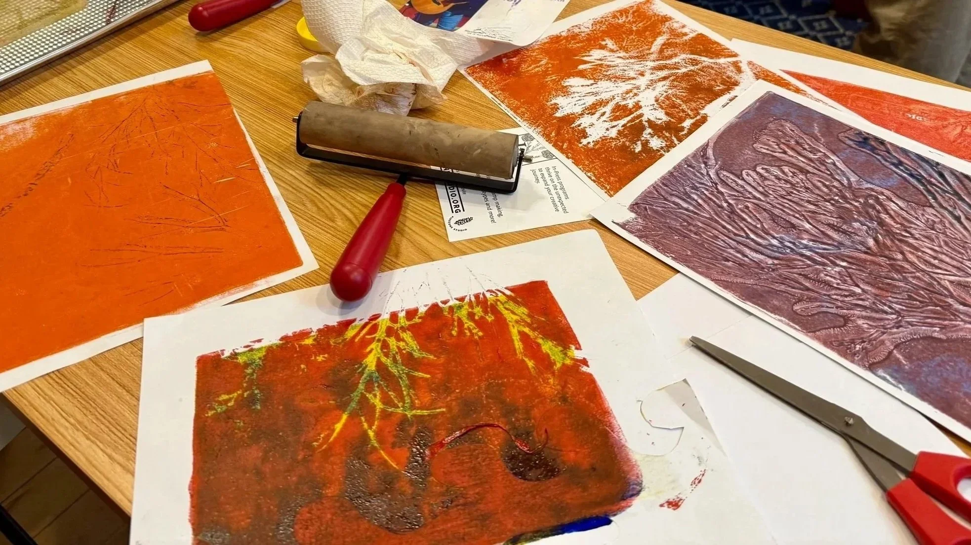 Artwork prints on paper with various textures and colors spread on a wooden table, alongside tools including a brayer, scissors, and crumpled tissues.