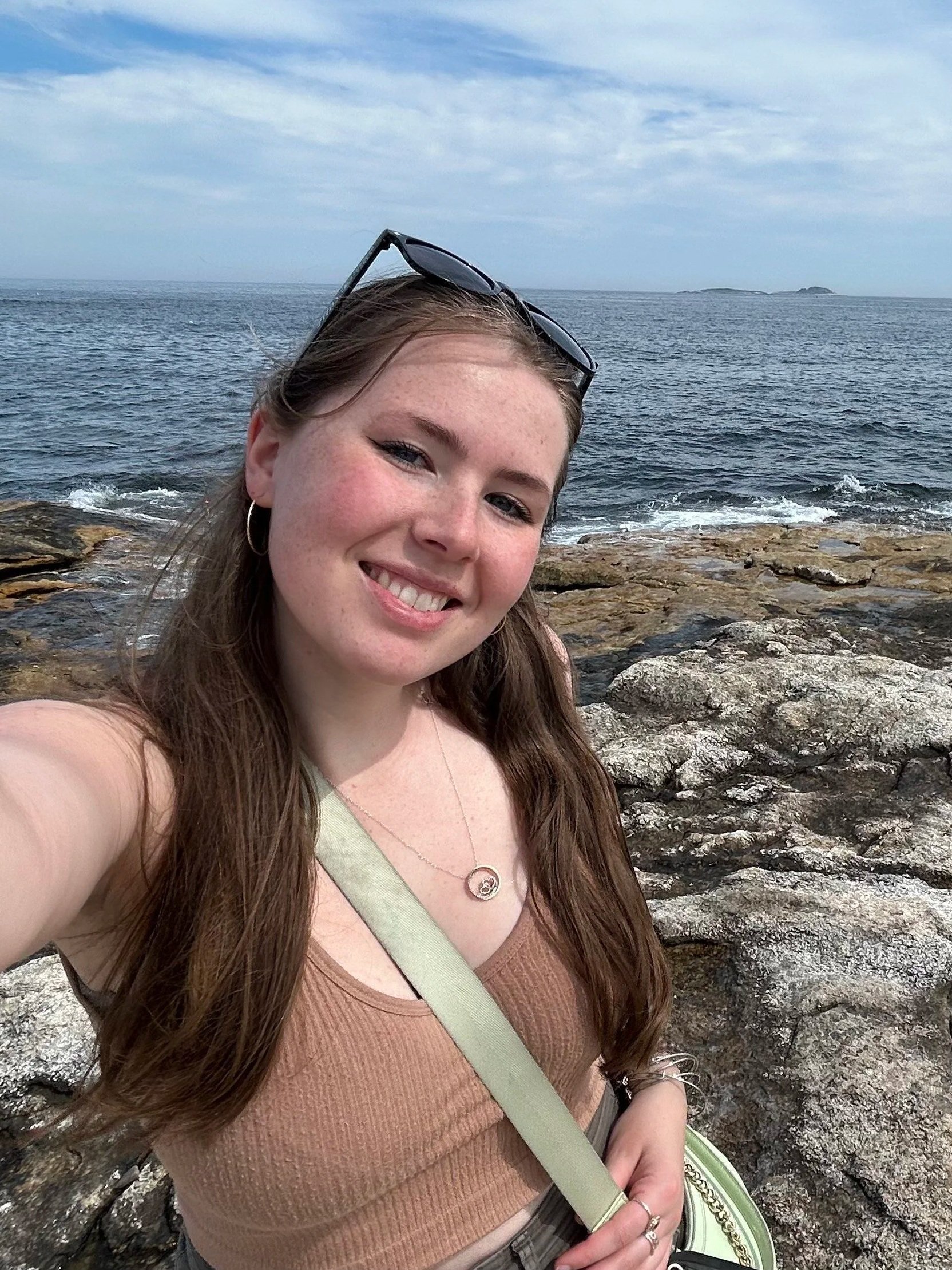 A young woman taking a selfie near the rocky shoreline with the ocean and islands in the background.