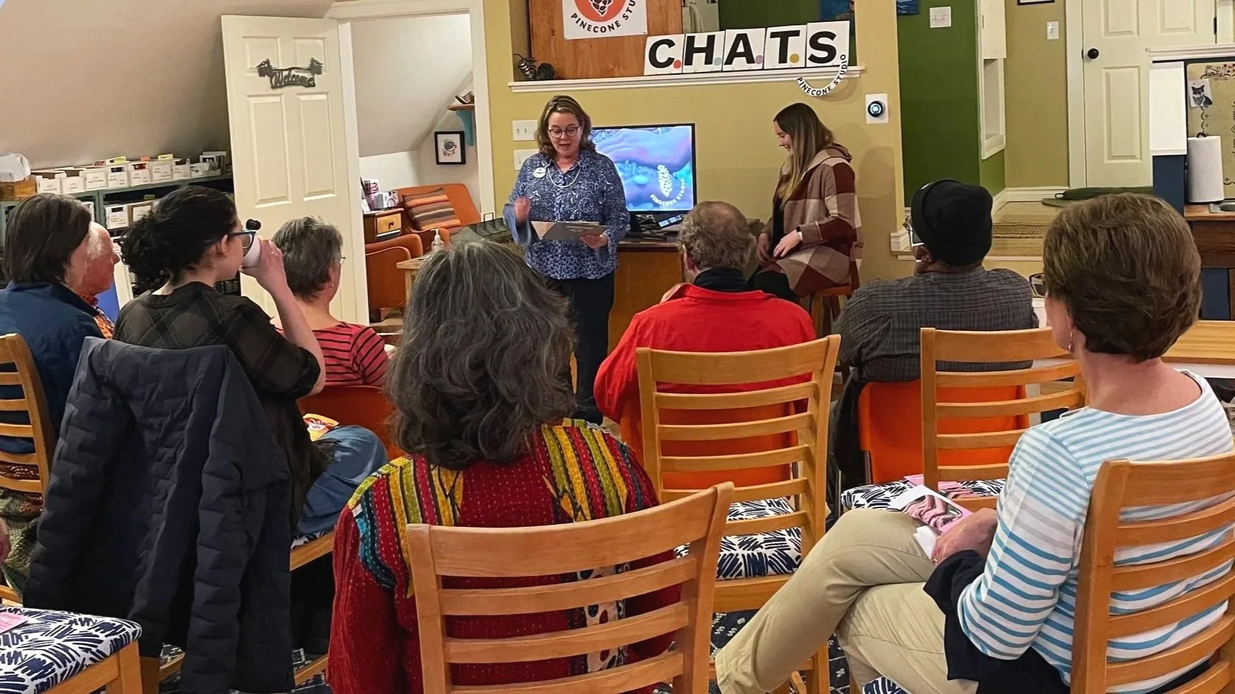Group of people sitting in a room listening to a woman speaking at a presentation table, with a sign that says 'Chats' and a television screen in the background.