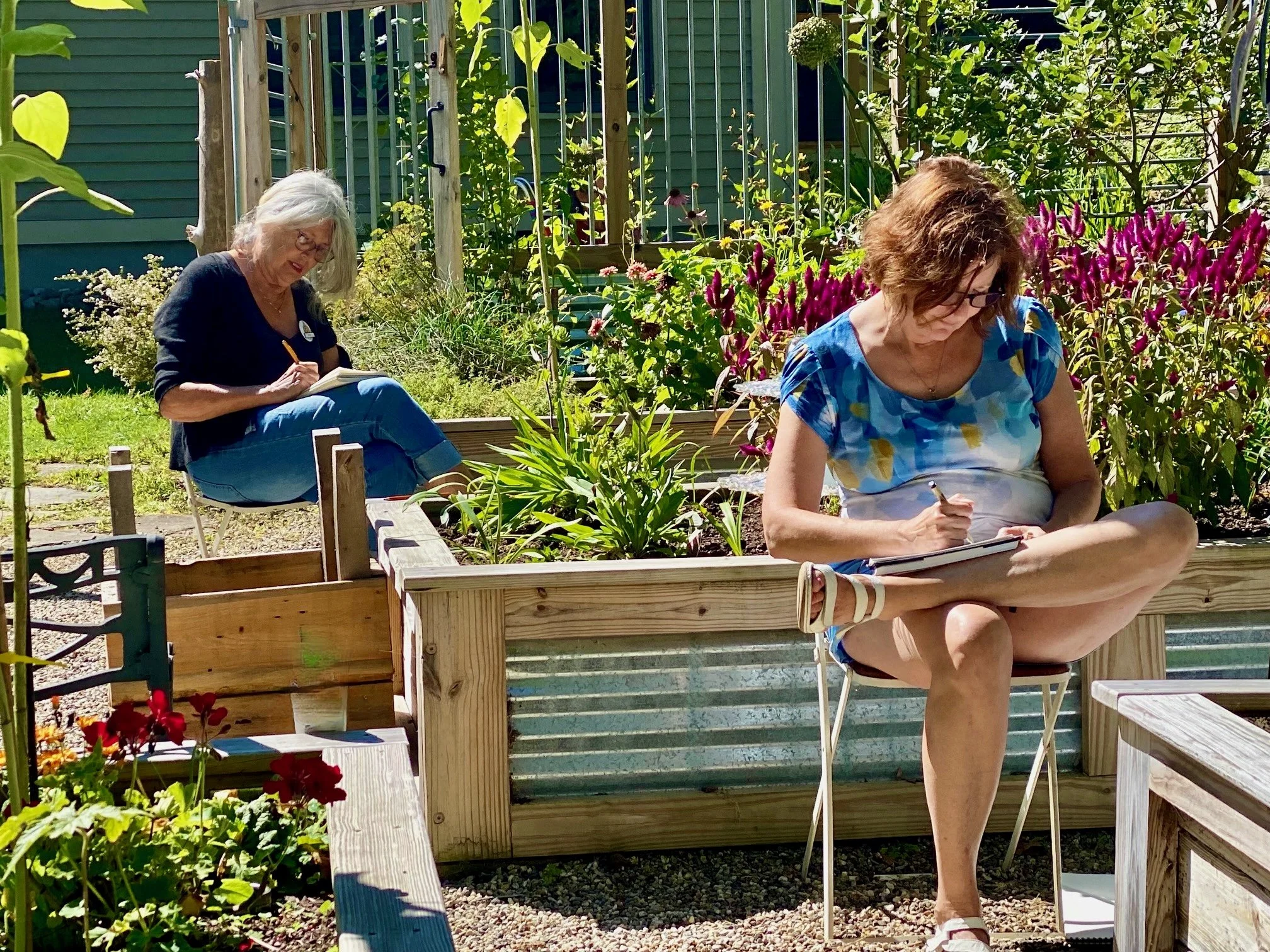 Two women sitting outdoors in a garden, sketching or drawing on paper. The woman in the foreground sits on a chair with one leg crossed over the other near a flower garden, while the woman in the background sits on a small chair surrounded by plants and flowers.