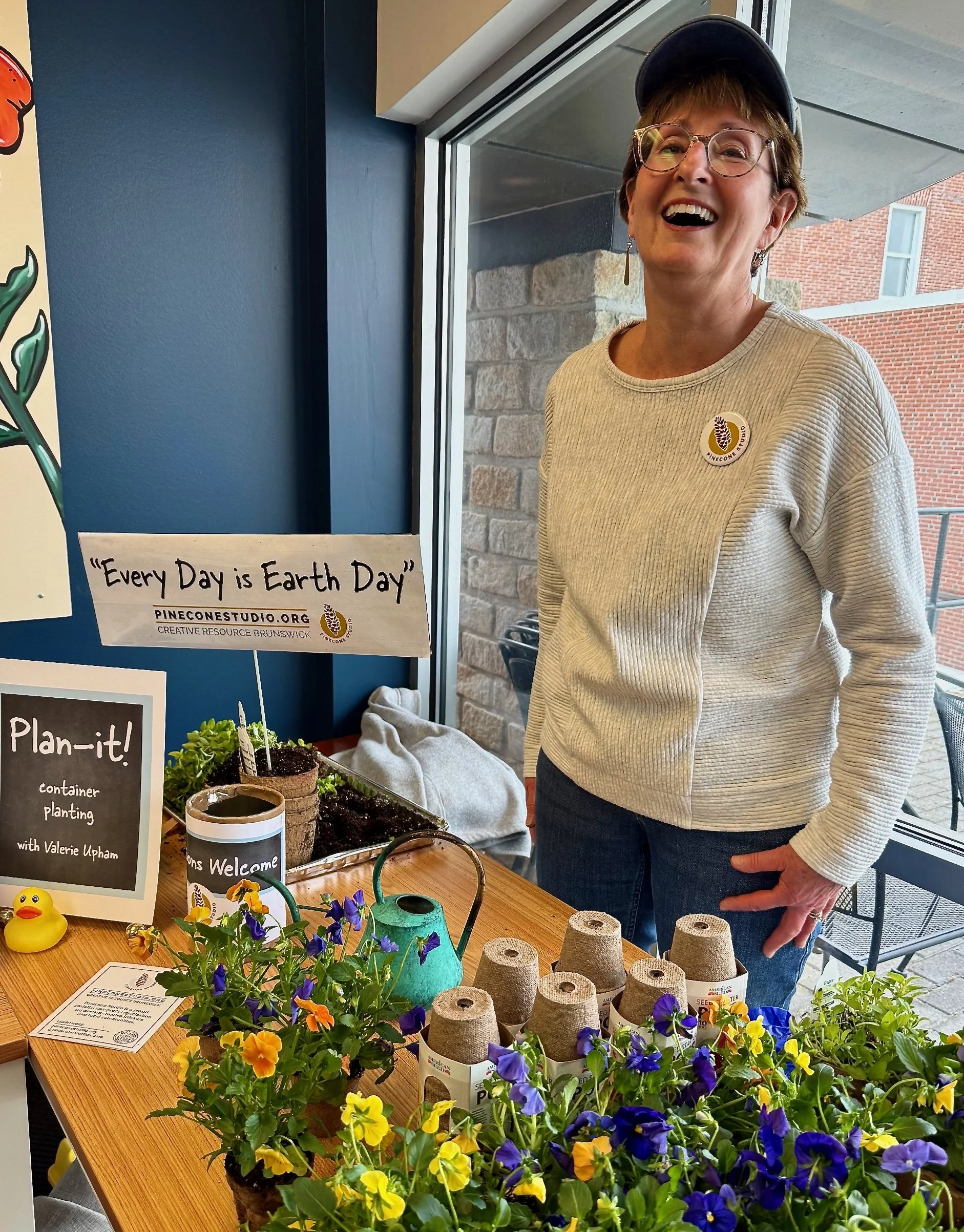 A smiling woman with glasses and short hair wearing a gray sweater and a blue cap standing behind a table with potted flowers, gardening supplies, and a sign that says 'Every Day is Earth Day'."