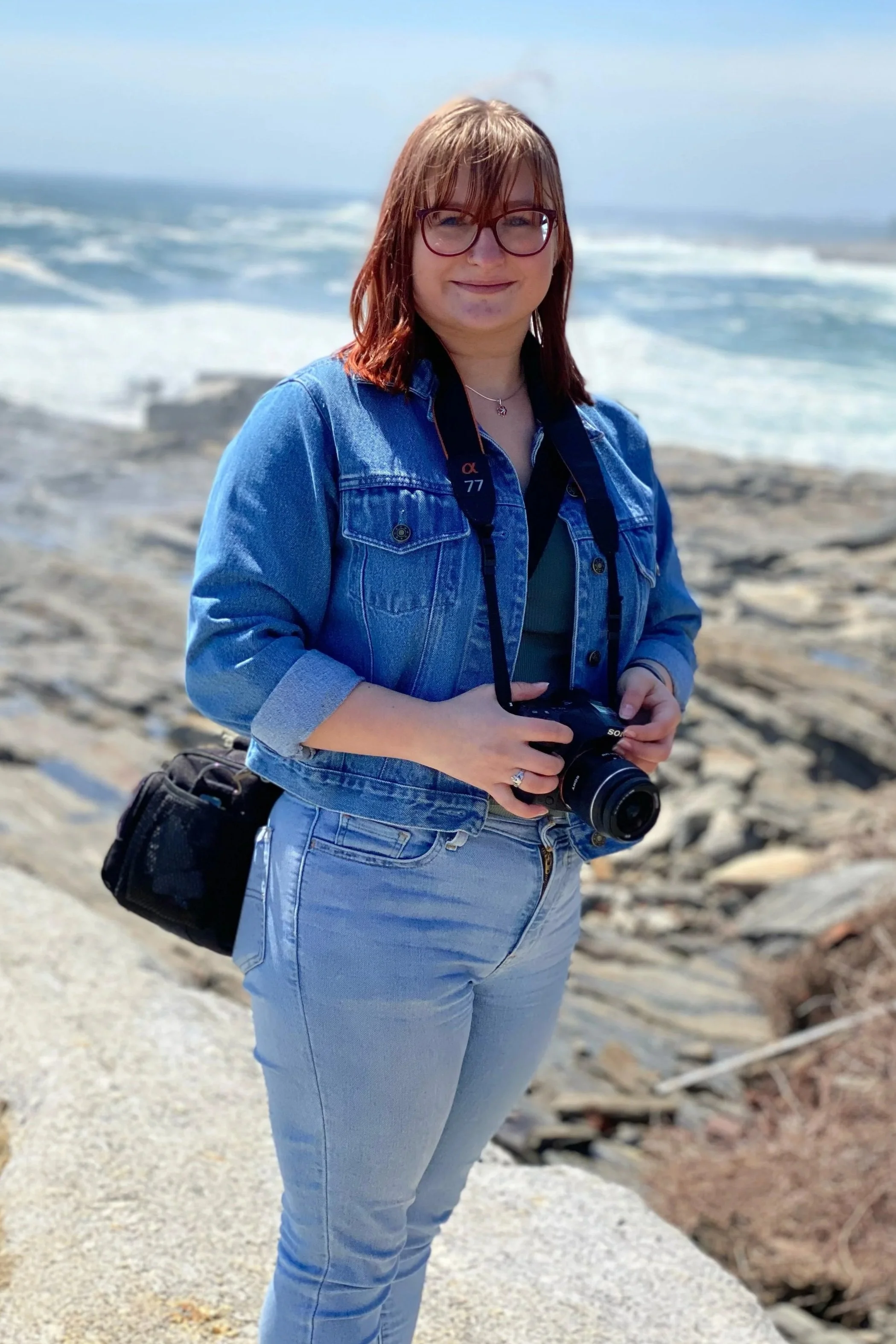 Woman holding a camera on a rocky beach with ocean waves in the background.