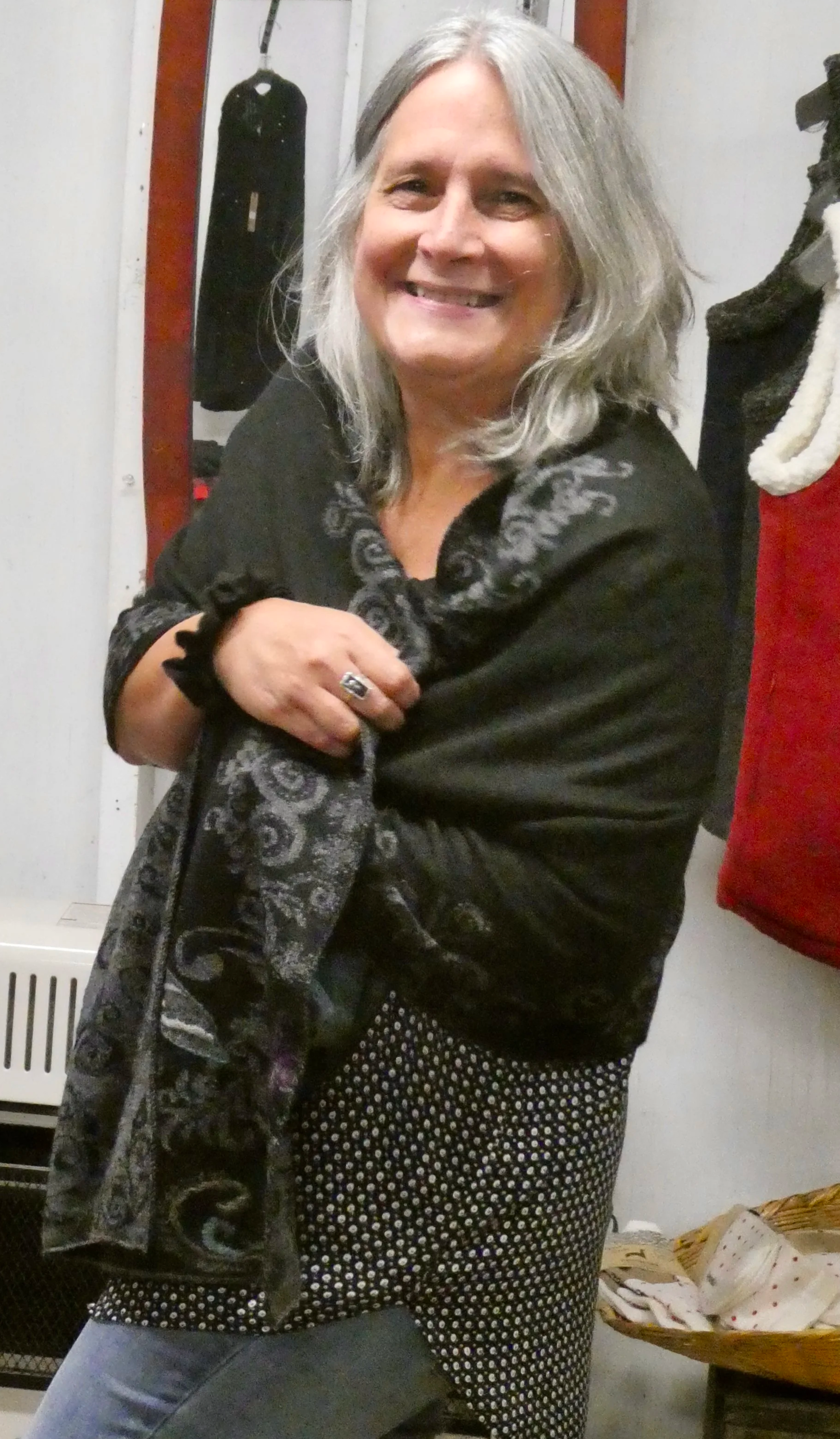 A middle-aged woman with shoulder-length gray hair smiling while wrapping a patterned black and gray scarf around her shoulders in a cozy, indoor setting.