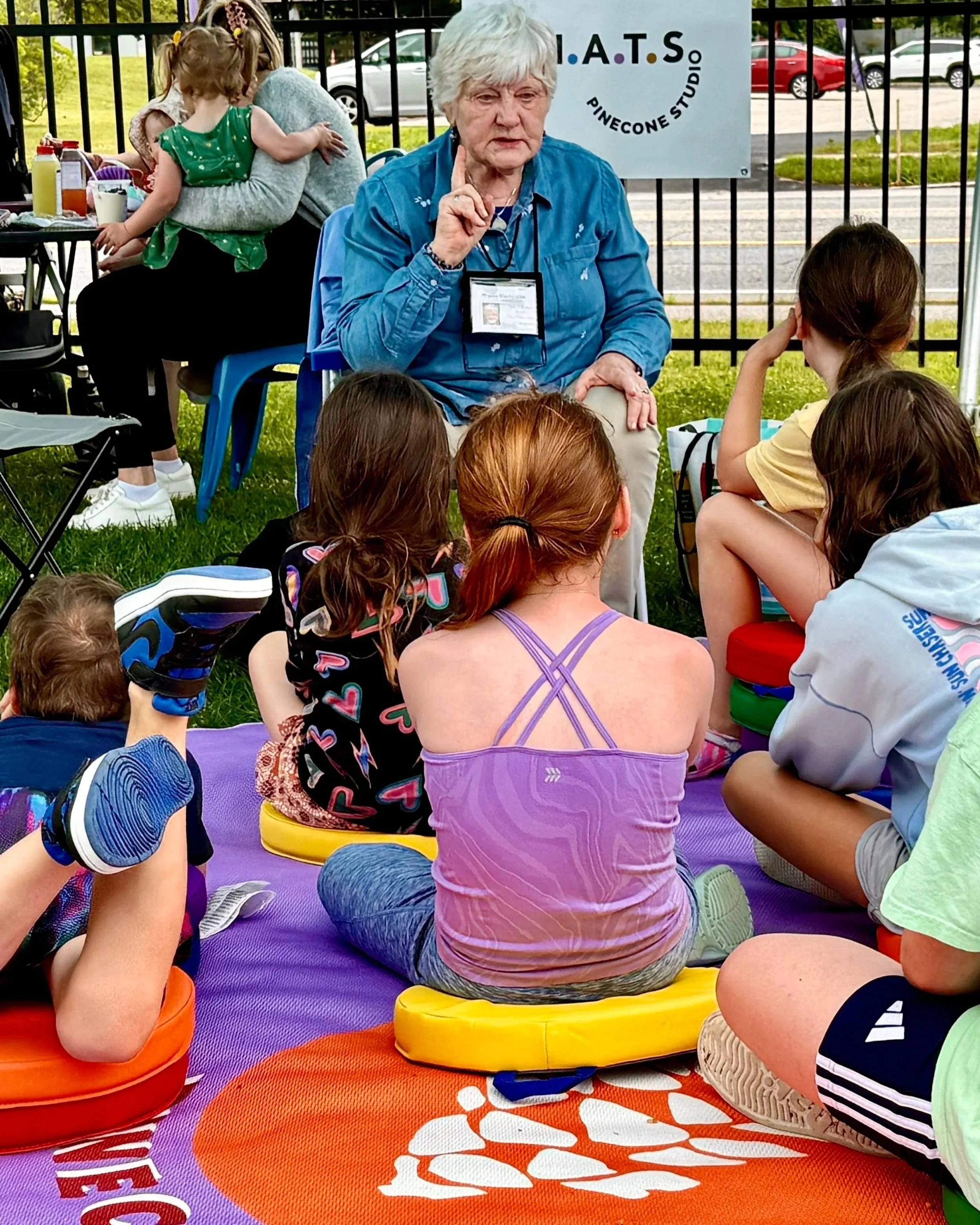 An elderly woman with white hair and a blue shirt is sitting outdoors, speaking or sharing with a group of children who are seated on colorful cushions on the grass. There is a sign behind her that reads 'I.A.T.S. Pinecone Studio,' and a woman with two children is in the background near a table with supplies.