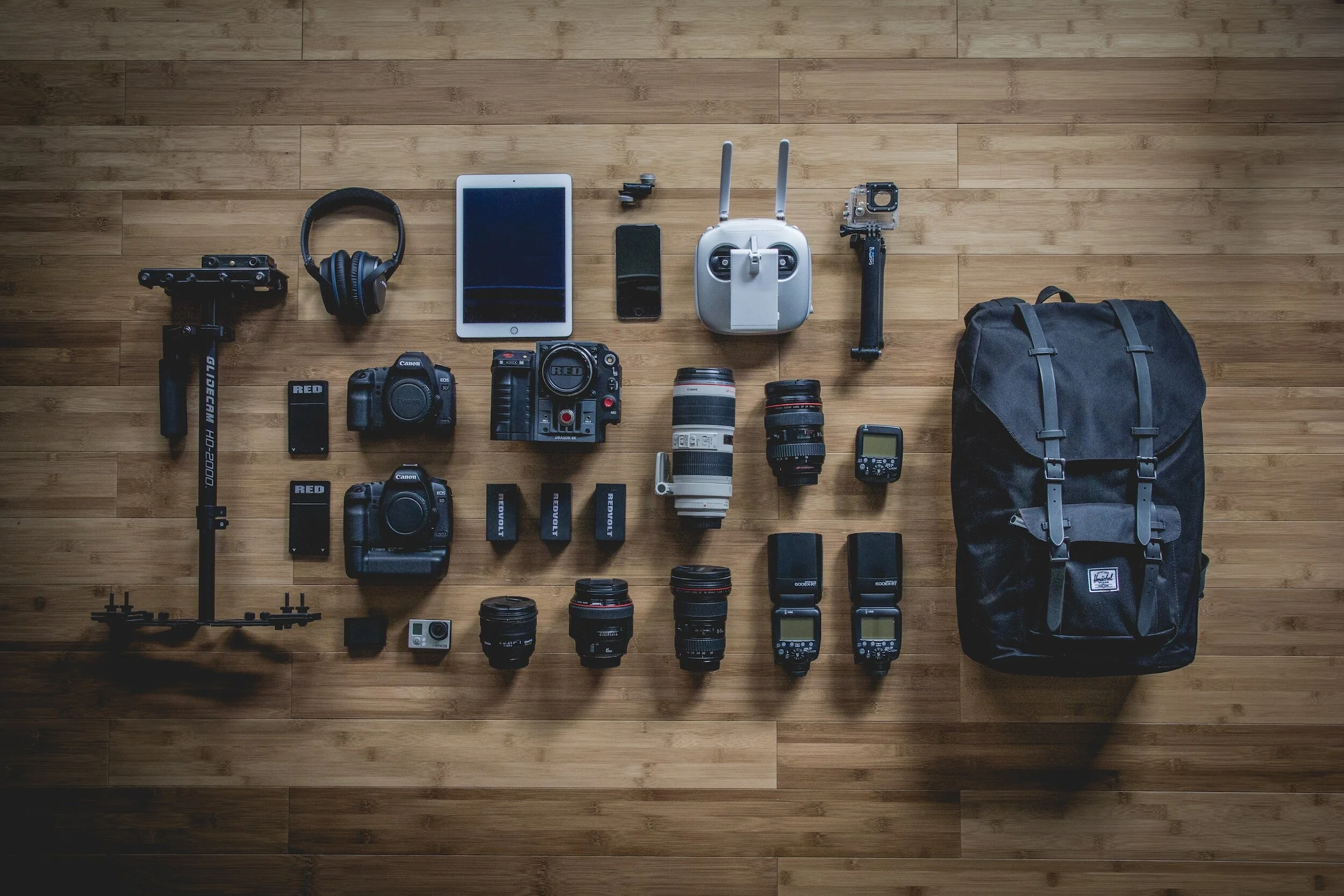 Flat lay of various camera equipment and accessories, including cameras, lenses, stabilizer, headphones, tablet, smartphone, drone controller, GoPro, speedlights, and a black backpack on a wooden surface.