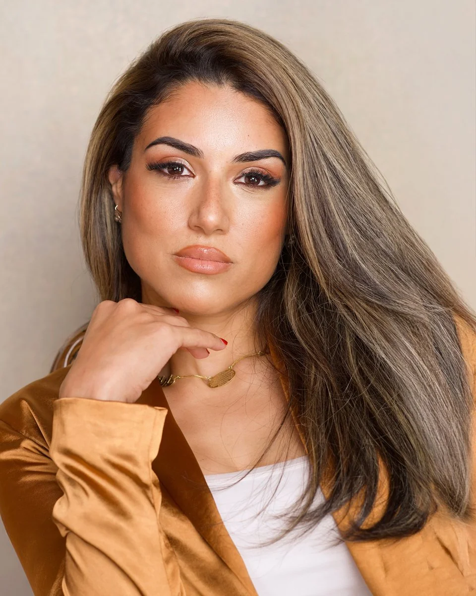 A woman with long, styled brown hair, wearing a white top, a brown satin jacket, and gold jewelry, posing with her hand near her chin against a plain background.