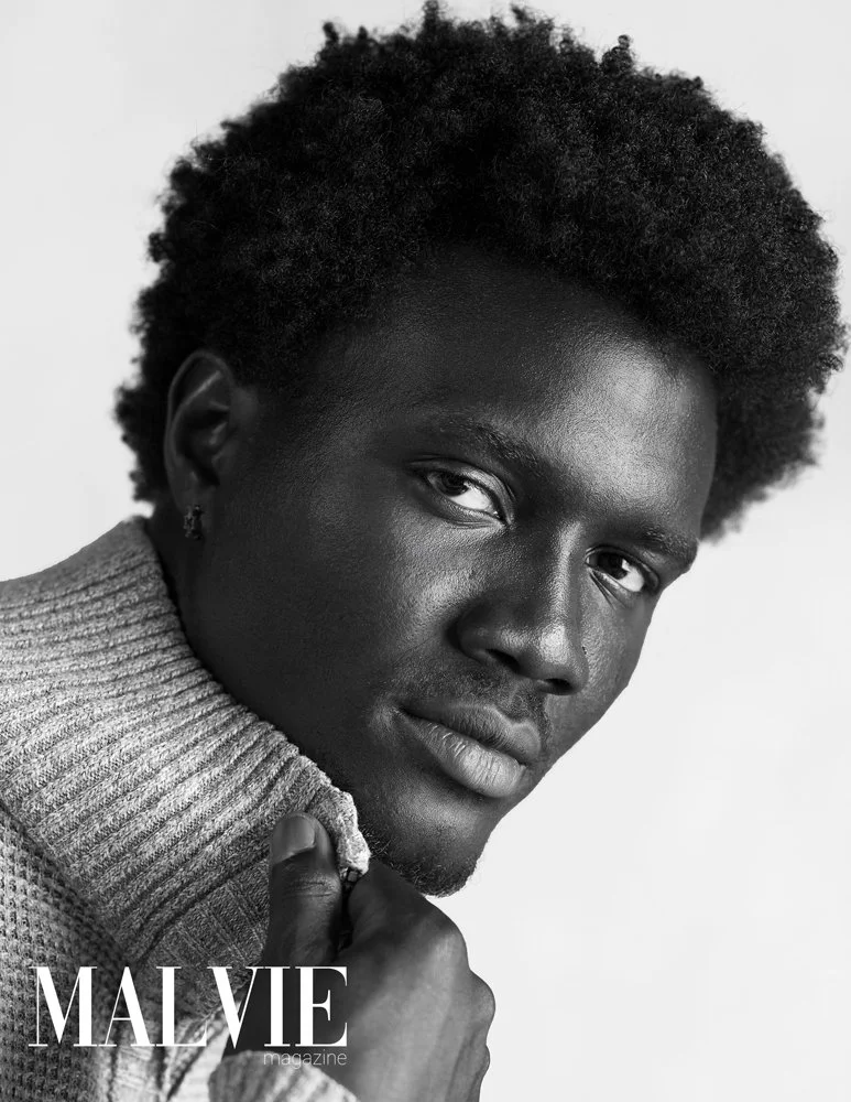 Black man with natural curly hair wearing a knitted turtleneck, gazing into the camera, for Malvie Magazine.