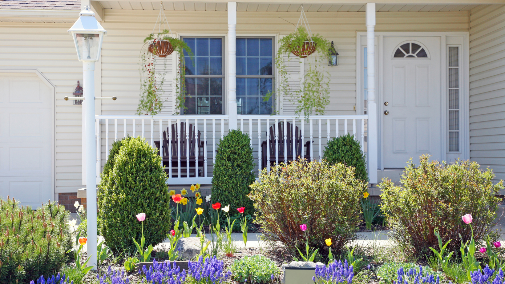 front yard of a White House with a white door and front patio, front yard has lots of flowers including tulips and lavender, bushes, and a lamppost, the patio has two chairs and two hanging plants, this is a home in the spring time