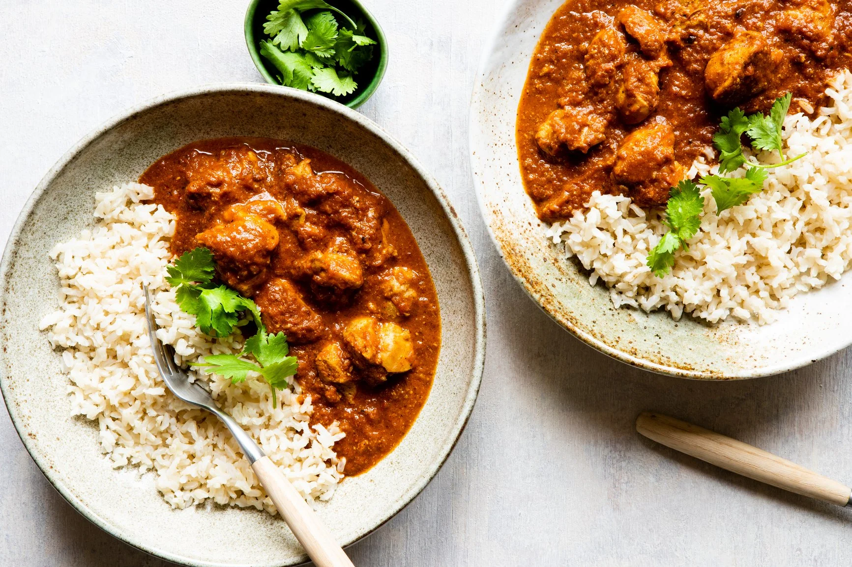 Two bowls of chicken curry served with white rice, garnished with cilantro, on a white surface with a small bowl of cilantro nearby.