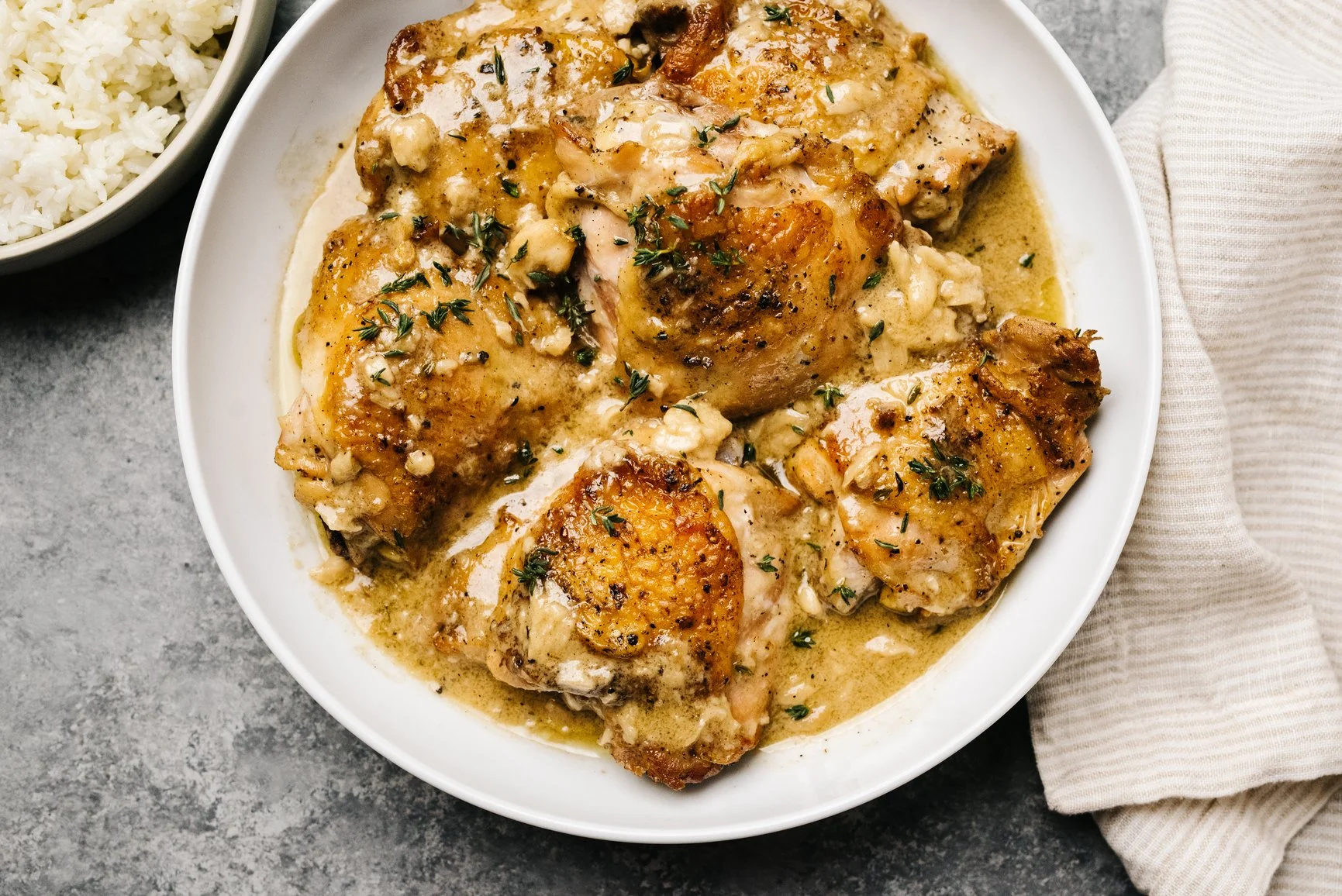 Baked chicken thighs in a creamy garlic sauce garnished with chopped herbs, served in a white bowl. A small bowl of rice is visible nearby, on a gray surface with a beige napkin.
