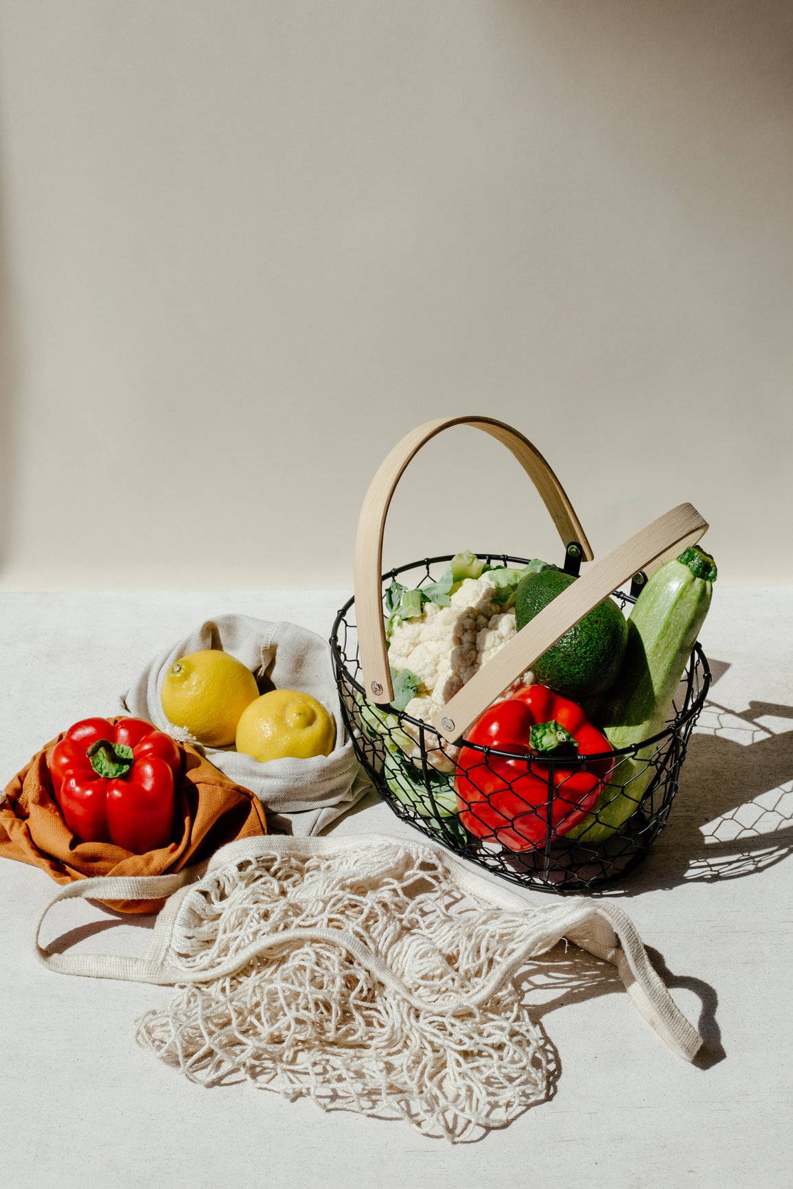 A basket of fresh vegetables and fruits on a white surface, with some produce outside the basket, including red and yellow bell peppers, also a cauliflower, zucchinis, and avocados, with a cloth and a net bag nearby.