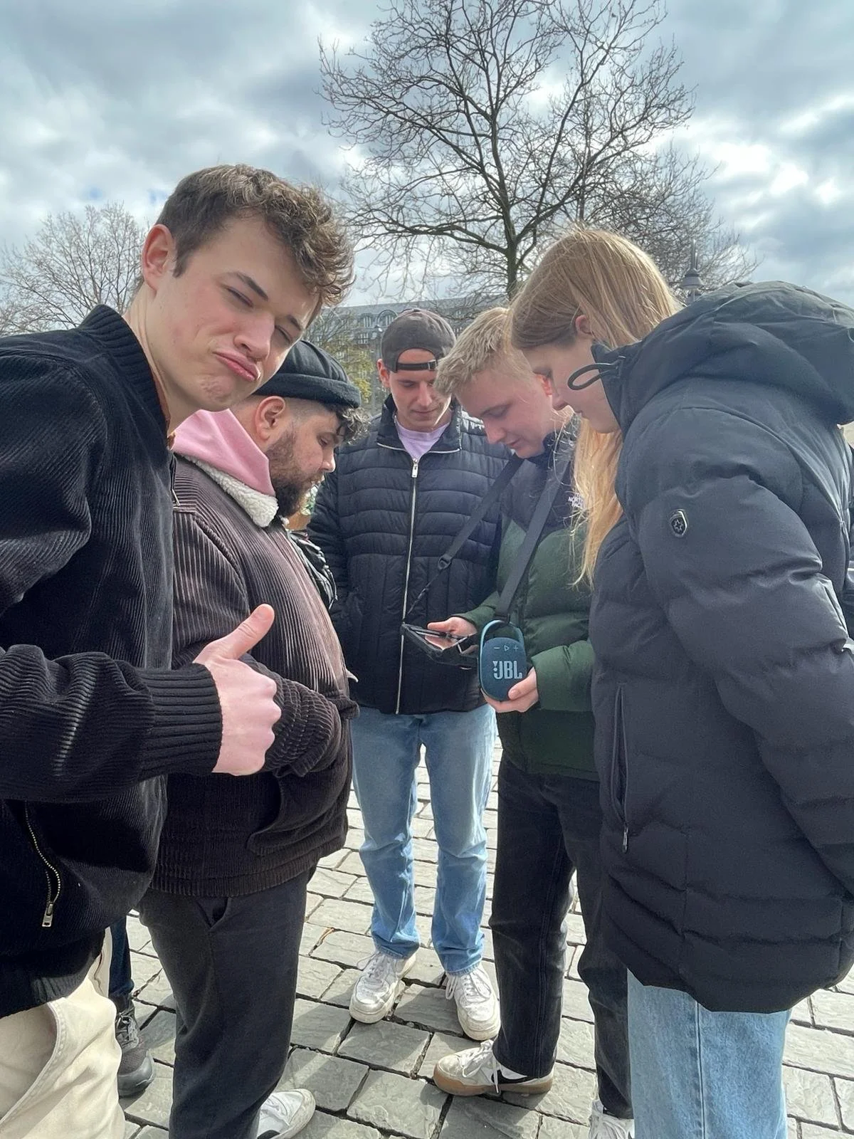 Eine Gruppe junger Menschen steht im Freien auf Kopfsteinpflaster und betrachtet ein Smartphone, während einige nach unten schauen.