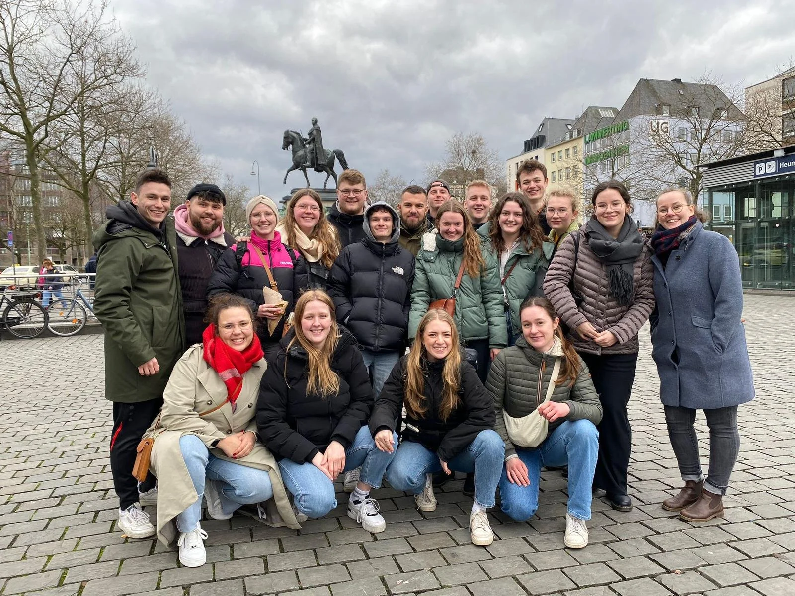 Gruppe junger Menschen vor einem Reiterstandbild in einer Stadt, im Hintergrund mehrstöckige Gebäude und Bäume, bewölkter Himmel.