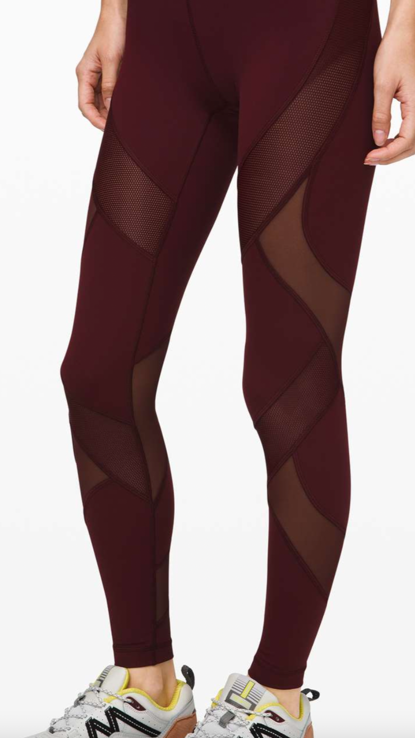 Close-up of a person wearing maroon activewear leggings with mesh and sheer panels and white athletic shoes, standing against a white background.