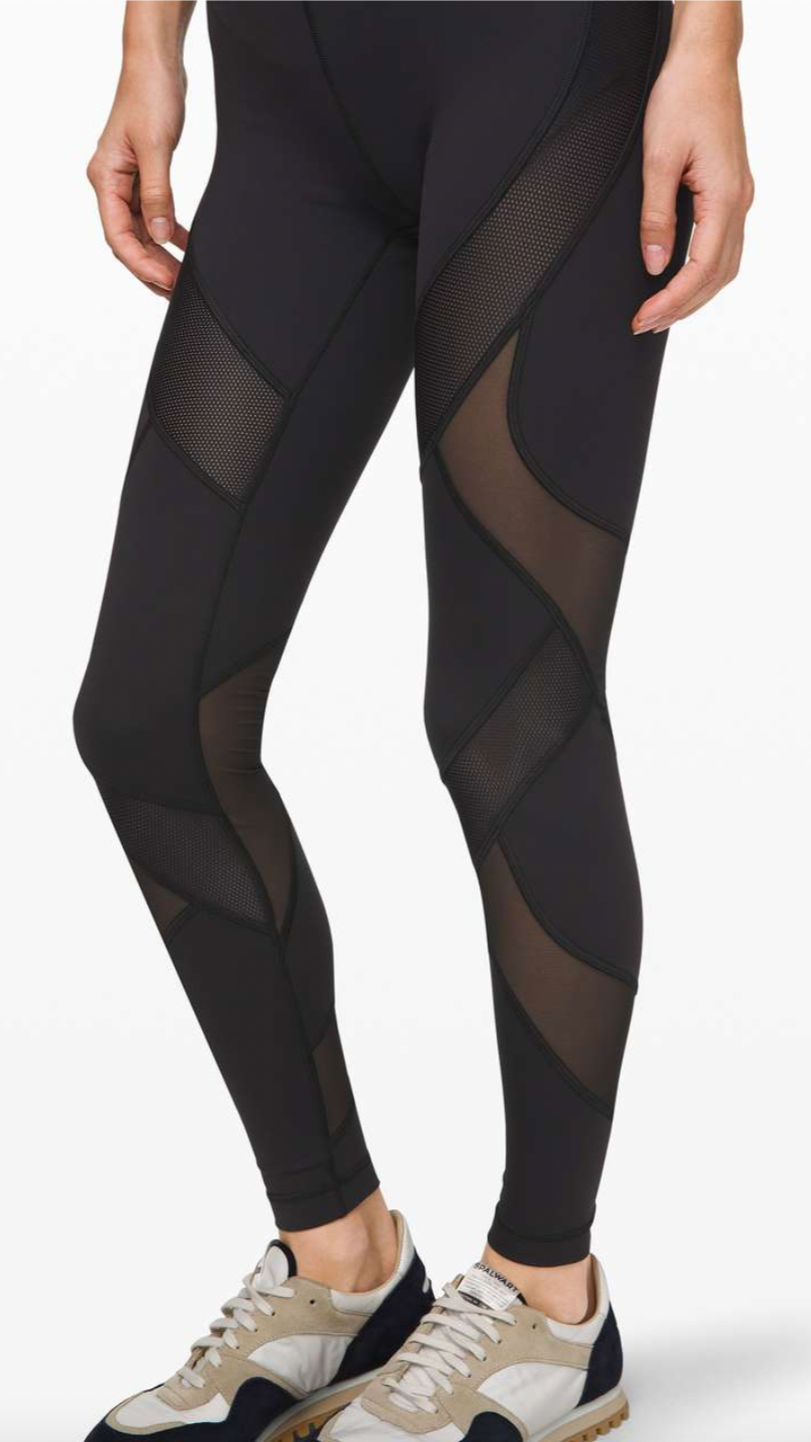 Close-up of a person wearing black workout leggings with mesh and sheer panels, standing with hands relaxed beside their hips, and wearing beige and black sneakers.