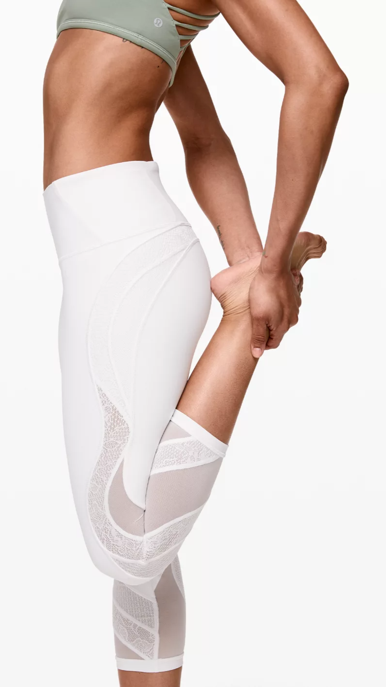 A person stretching and holding their foot, wearing white athletic leggings with mesh panels and a light green sports bra with cutouts.