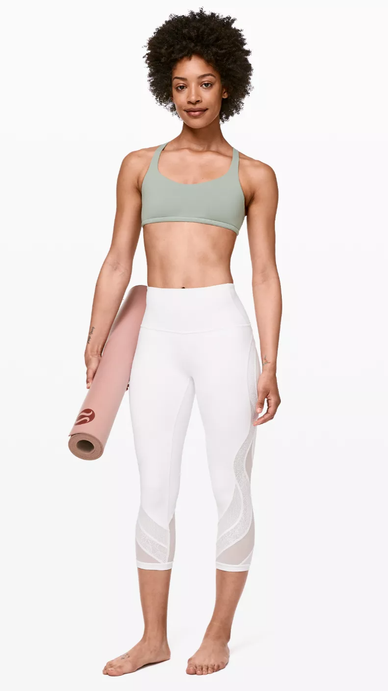 A woman with curly hair holding a yoga mat, wearing a light green sports bra and white leggings with mesh panels, standing barefoot against a white background.
