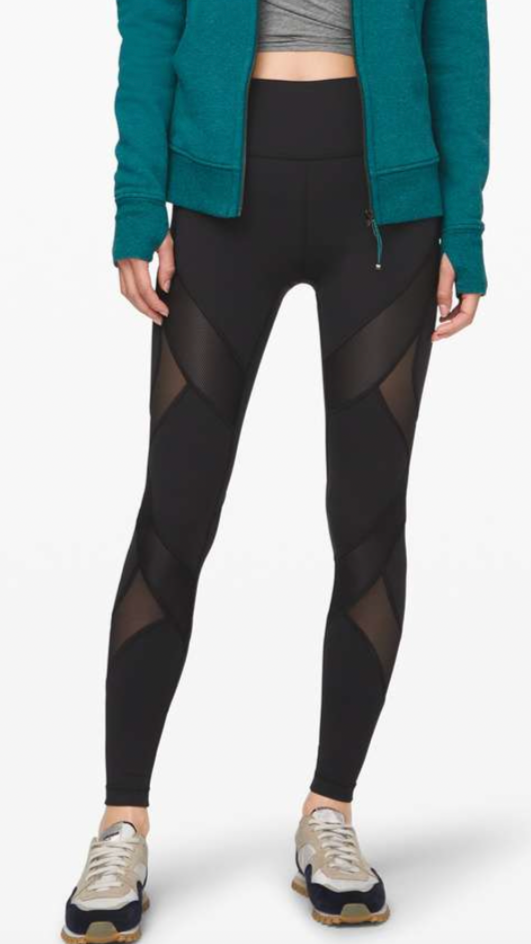 A woman in athletic wear standing against a plain white background, wearing a teal zip-up hoodie, gray crop top, black patterned leggings, and sneakers.