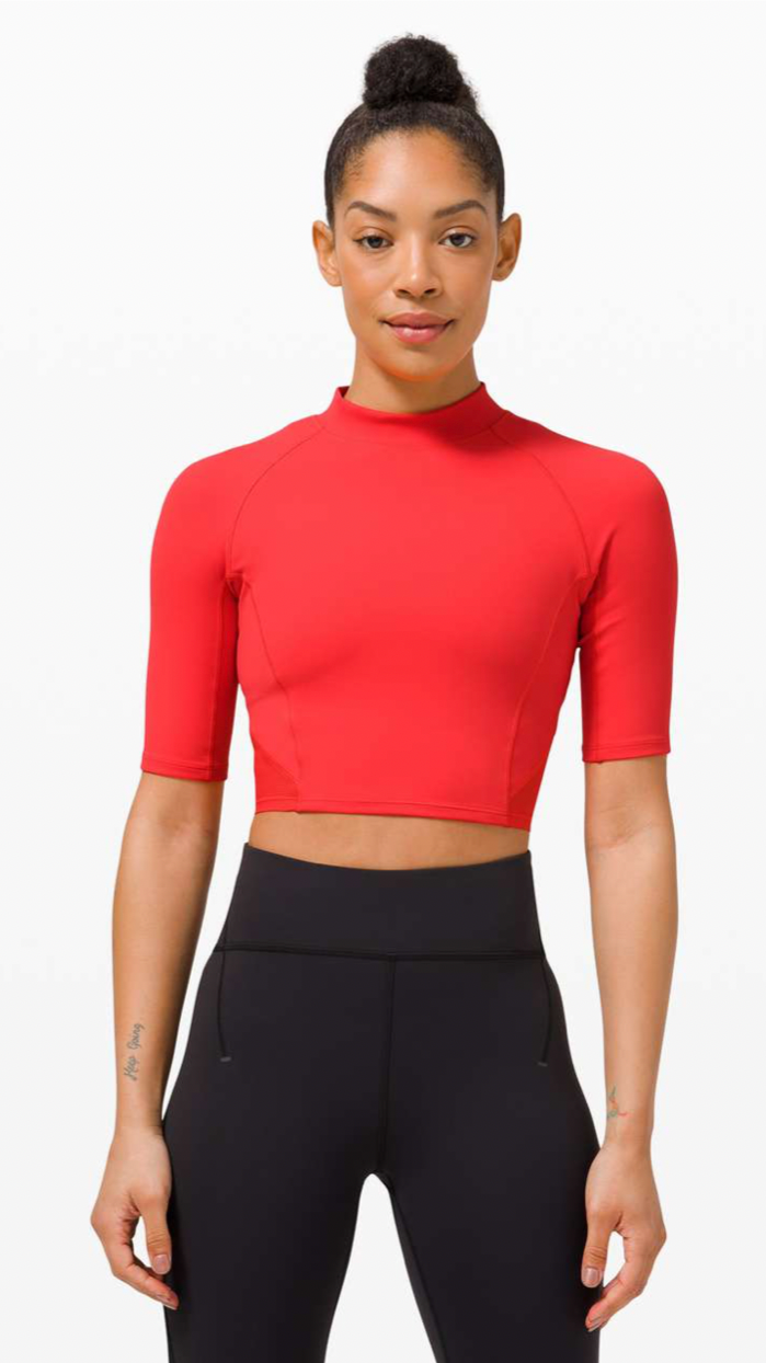 A young woman with dark hair in a bun, wearing a red short-sleeve athletic crop top and black high-waisted leggings, standing against a plain white background.