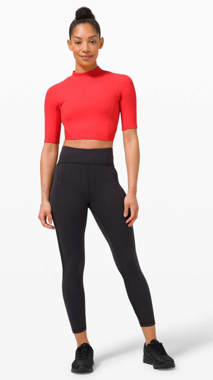A woman standing in athletic wear, wearing a red crop top, black leggings, and black sneakers in front of a plain white background.
