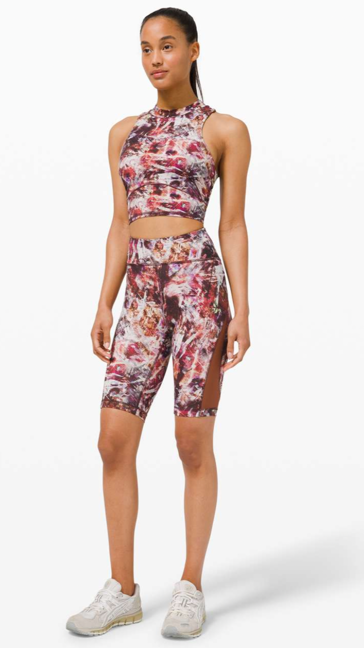 Young woman wearing colorful patterned athletic crop top and matching shorts with side slits, standing against a white background.