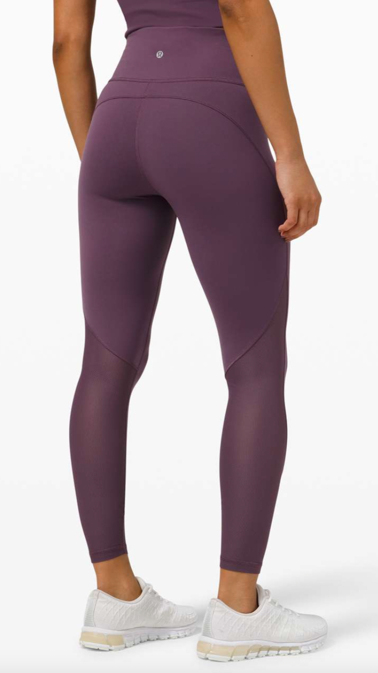 Woman standing wearing purple high-waisted leggings and white sneakers against a white background.