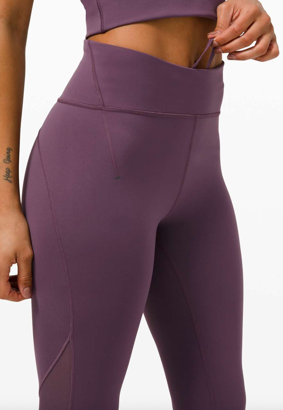 Close-up of a woman in purple athletic leggings and sports bra, with her hand pulling the waistband of her leggings to reveal a hidden pocket.
