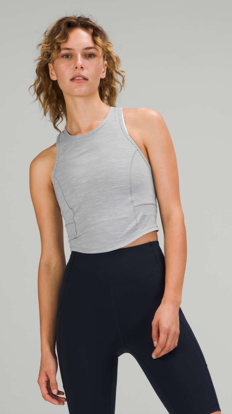 A woman in gray athletic tank top and black workout leggings stands against a plain background.