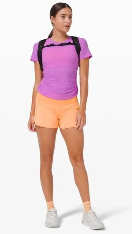 A young woman wearing a purple t-shirt, peach shorts, white sneakers, and a black backpack, standing against a white background.