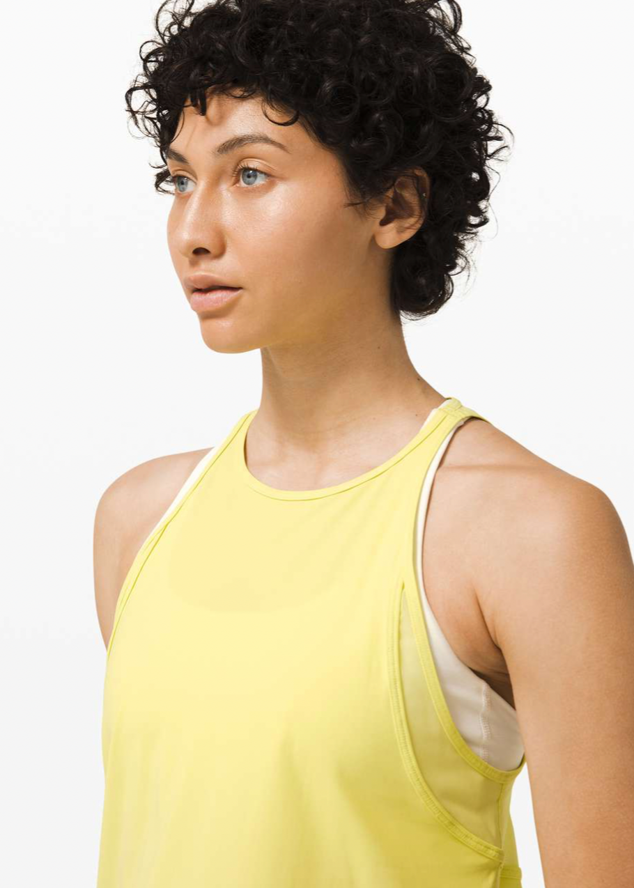 A woman with short curly dark hair wearing a yellow sleeveless athletic top, looking to her left, with a plain white background.