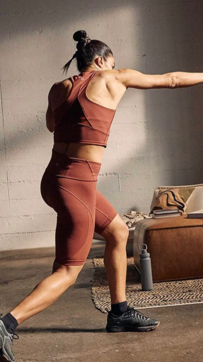 A woman doing a stretching exercise in a gym or workout space, wearing a matching brown sports bra and shorts, black shoes, and black socks, with a water bottle and books on a brown ottoman nearby.