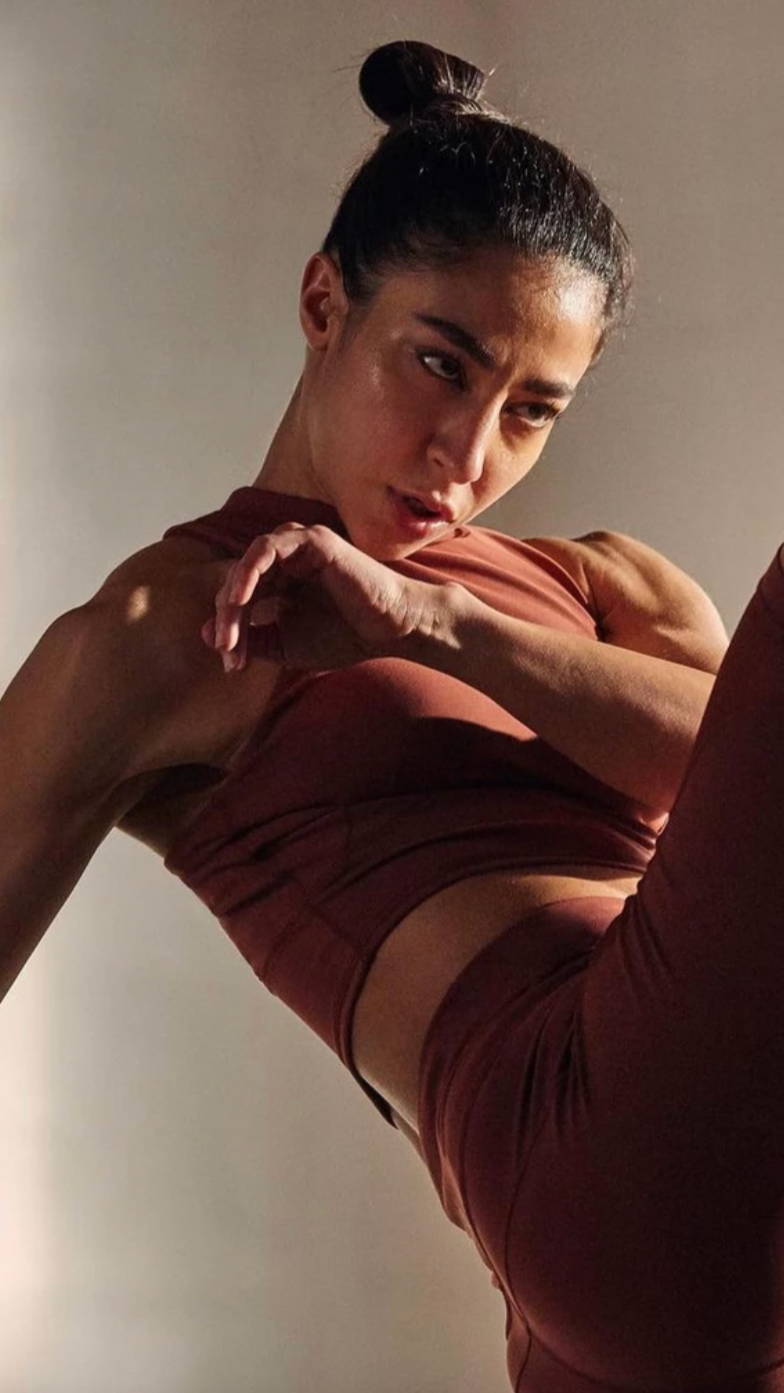A woman in athletic wear is performing a fitness move on a plain wall background.