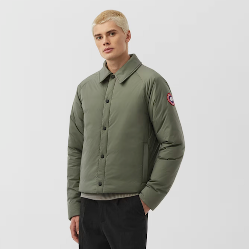 Young man with platinum blonde hair wearing a green quilted jacket with a logo patch on the sleeve, standing against a plain background.