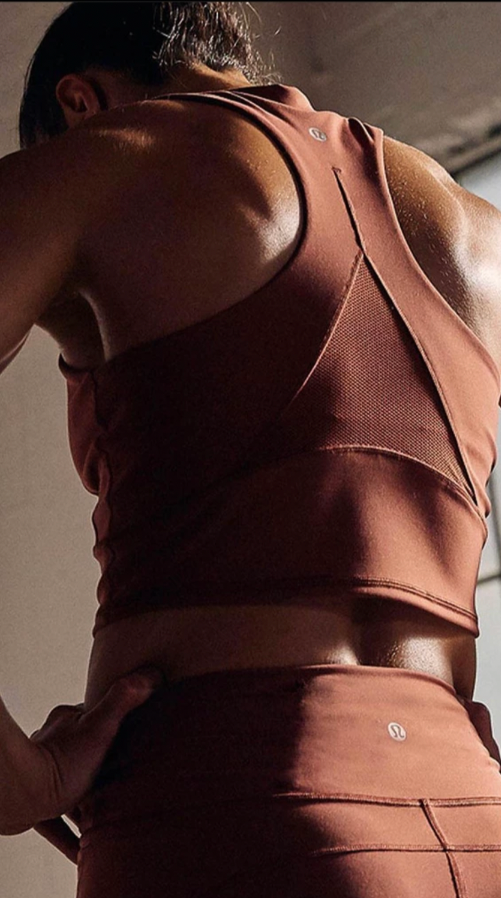 A woman wearing a matching rust-colored athletic sports bra and leggings taking a mirror selfie, with her hand on her hip. The background shows a bedroom with a bed and pillows.