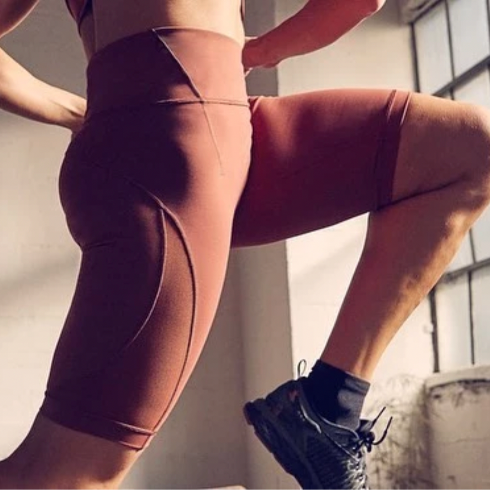 A woman in athletic wear stretching in a room with large front windows, wearing black running shoes and brown leggings.