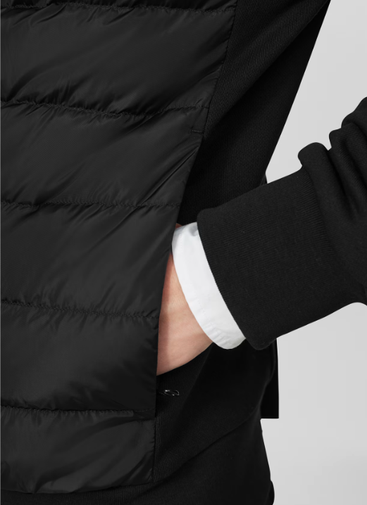 Close-up of a person wearing a black puffer vest over a black sweatshirt with a white shirt underneath, with one hand in the pocket.