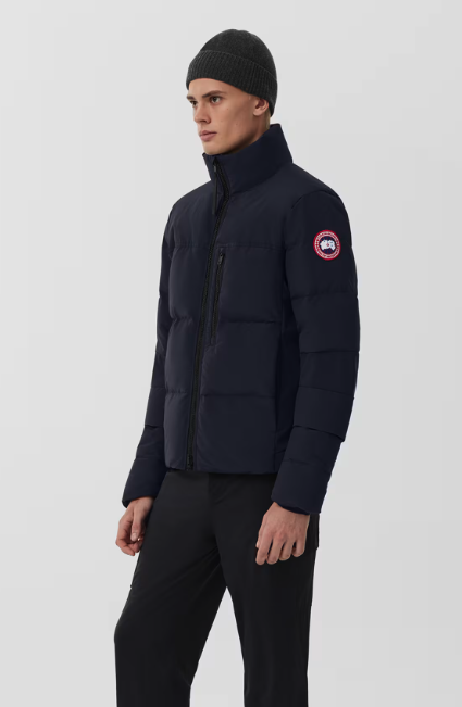 Man wearing a dark puffer jacket with a logo patch on the sleeve and a gray knit beanie, standing against a plain light background.