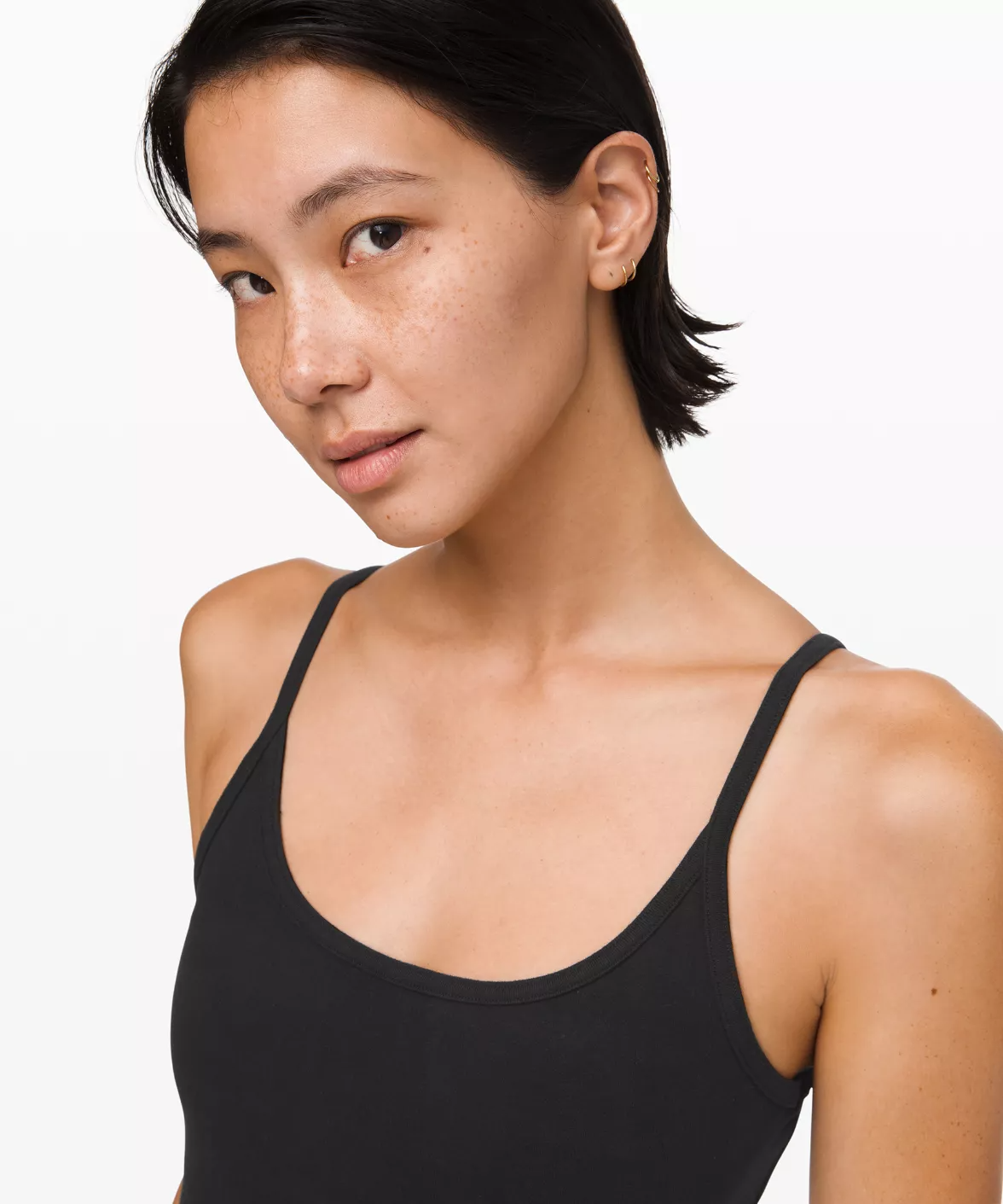 A woman with short black hair, light brown skin, and freckles, wearing a black tank top, posing against a plain white background.