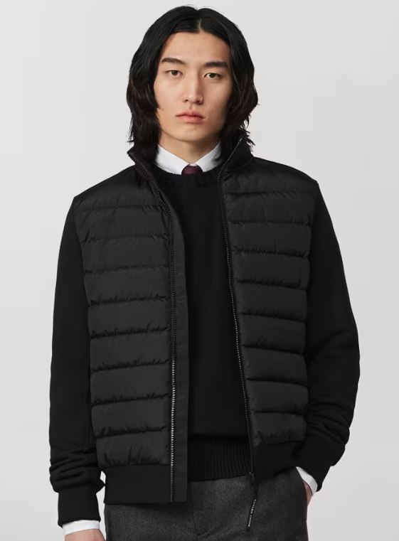 Young man with shoulder-length black hair wearing a black puffer vest over a black sweater with a white shirt and a maroon tie, standing against a plain light gray background.