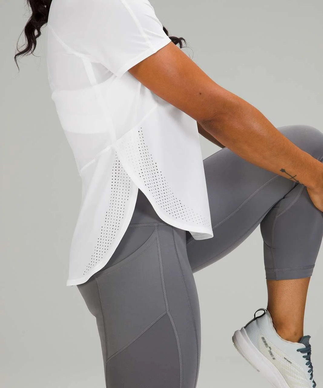 Person wearing a white athletic shirt, gray leggings, and white running shoes, stretching indoors.
