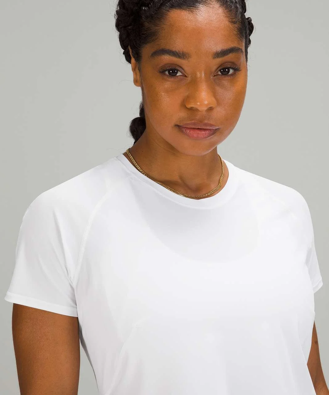 A woman with braided hair wears a white athletic shirt and a gold necklace, posing against a neutral background.