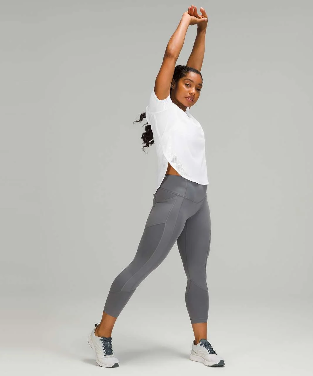 Woman in gray athletic leggings and a white short-sleeve shirt stretching with arms above head on a plain gray background.