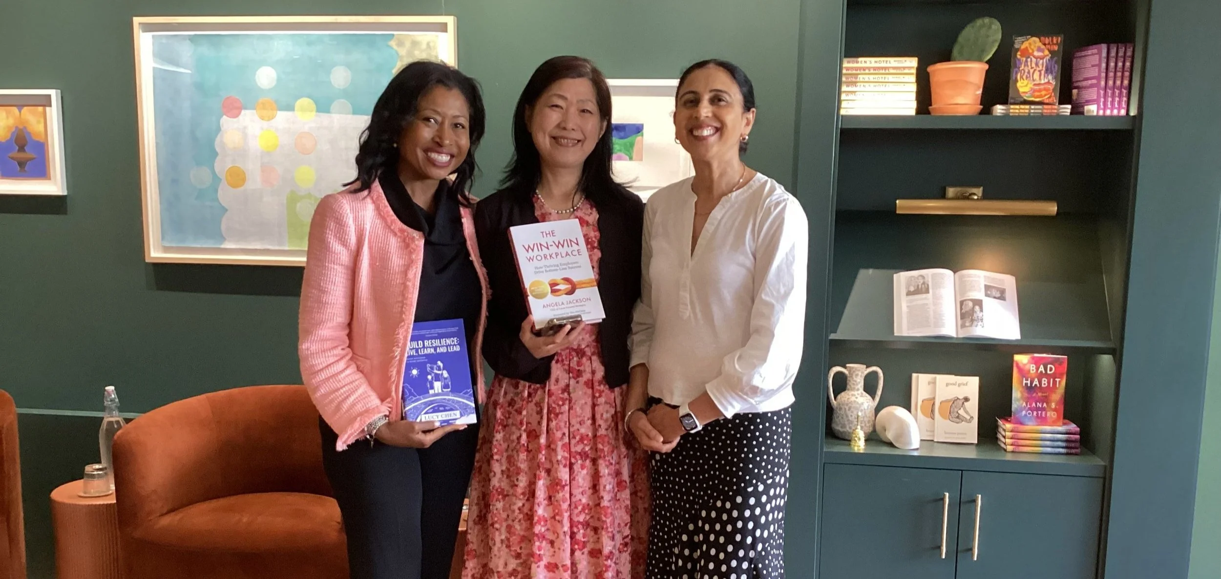 Three women standing together in a room with green walls and bookshelves, holding books and smiling at the camera.