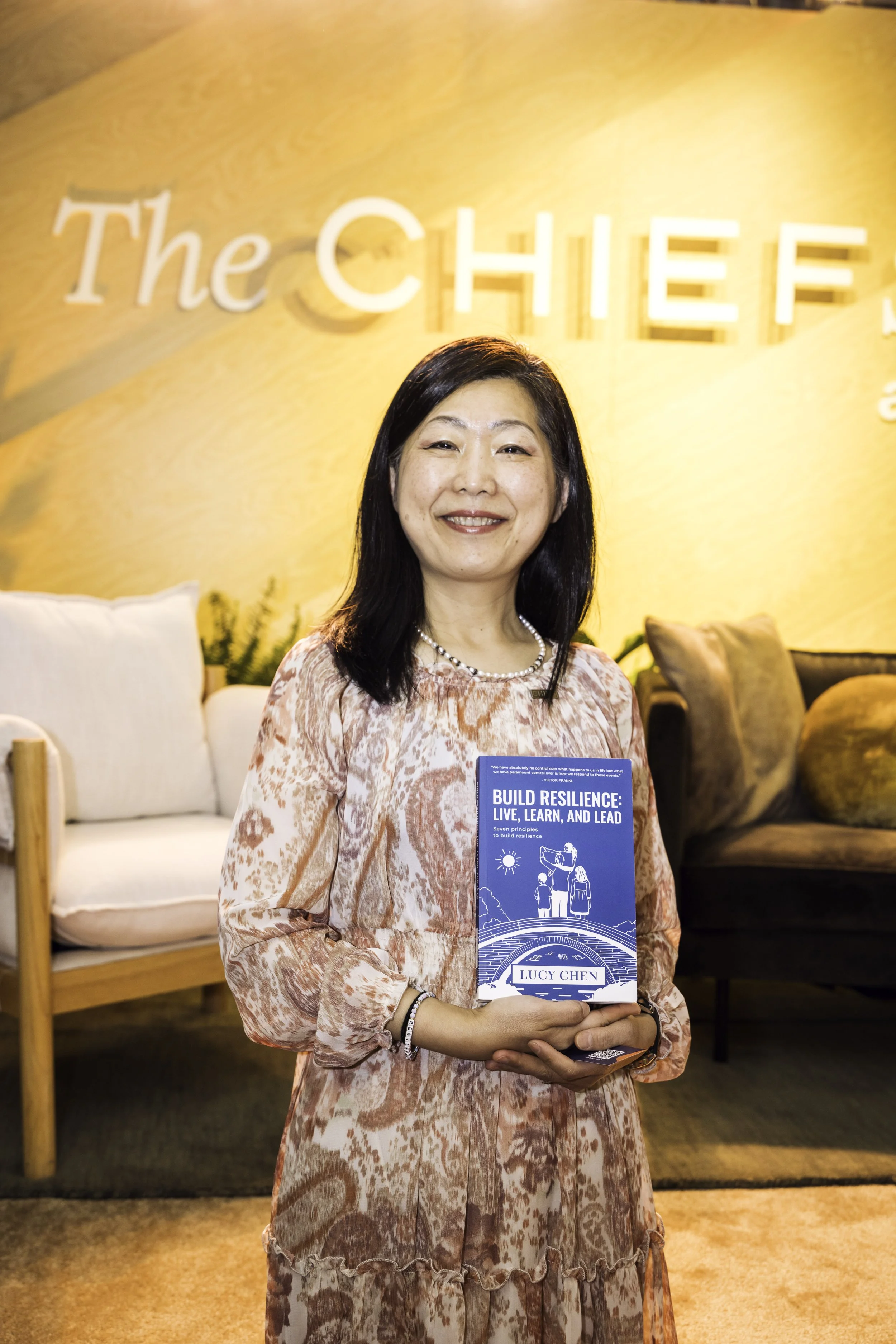 A woman standing indoors holding a book titled "Build Resilience: Live, Learn, and Lead" by Lucy Chen. She has shoulder-length black hair, wears a patterned dress, a pearl necklace, and a bracelet. Behind her is a beige couch, a dark couch, and a yellow-lit wall with partially visible text that reads "The CHIEF."