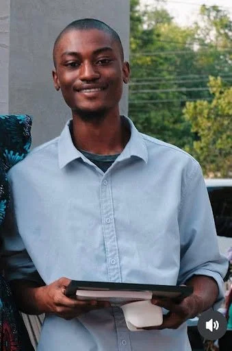 Medium shot of Fofanah standing and smiling in a blue button-down shirt. He holds his certificate and a book flat in his hands.